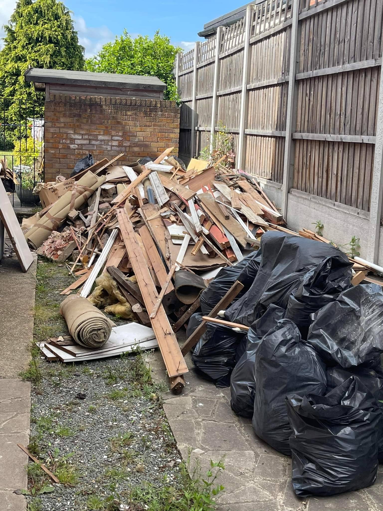 A pile of garden rubbish, including bin bags, leaves, branches, grass cuttings, and old wooden planks, stacked near a fence, ready for collection.